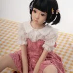 Realistic sex doll with anime-inspired features, young girl design, wearing a red and white gingham dress with lace collar, posing in a minimalistic setting with cherries on checkered fabric.