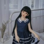 Realistic sex doll with long black hair in a casual bedroom setting, dressed in a navy blue dress with white stripes, designed for companionship and adult use.