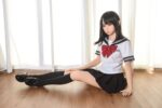 Realistic female adult doll with anime-style appearance, wearing a Japanese schoolgirl uniform, sitting on wooden floor in front of sheer white curtains, designed for adult companionship and simulation.