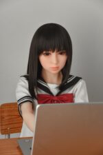 Realistic sex doll in schoolgirl outfit sitting at desk, focused on laptop, clear skin, detailed facial features, lifelike hair, close-up shot for adult companionship product imagery.