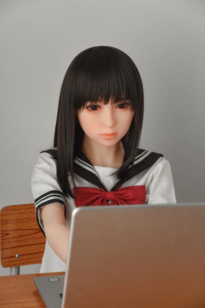 Realistic Sex Doll In Schoolgirl Outfit Sitting At Desk, Focused On Laptop, Clear Skin, Detailed Facial Features, Lifelike Hair, Close-Up Shot For Adult Companionship Product Imagery.