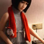 Bedroom girl with black bob wig and grey knit dress, red plush scarf, and white gloves, sitting at a black piano with electric outlet in background, indoor scene for realistic doll photography.
