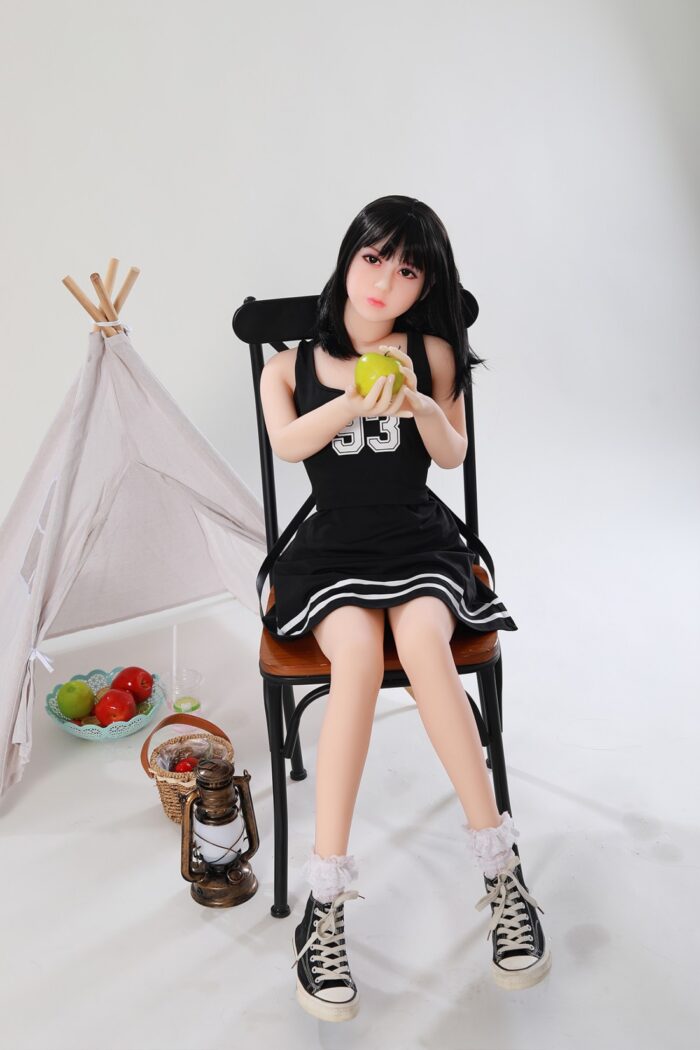 Elutruude näojoontega silikoonist seksinukk, mis istub toolil, hoiab käes rohelist õuna ning mille realistlik disain pakub intiimset seltskonda ja mugavust.