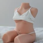 Cute sex doll with realistic body parts, dressed in a white tank top and thong, kneeling on a soft bed to showcase its detailed features and lifelike appearance.