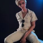 Realistična ženska figura seks lutke v ležerni kaki obleki s kratkimi svetlimi lasmi, ki sedi na stolčku na temnem ozadju z modro osvetlitvijo. Popolna za užitek odraslih in zbirateljski eksponat.