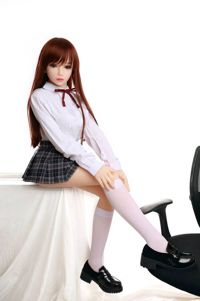 Realistic Sex Doll In Schoolgirl Outfit With Long Auburn Hair, White Blouse And Plaid Skirt, Posed Sitting On A Surface With One Leg Raised, Highlighting Its Detailed Mannequin Design And Lifelike Features.