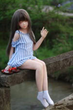 Realistic sex doll girl sitting on a stone bridge outdoors, wearing a blue and white striped dress, white socks, in a natural green environment, showcasing detailed craftsmanship.