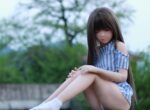 Realistic sex doll in outdoor setting, soft focus, detailed body features, with blue and white striped dress, long brown hair, and closed eyes, outdoor greenery background, ideal for intimate companionship images.