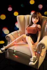 Realistic sex doll sitting on a beige armchair decorated with colorful string lights, with blurred colorful bokeh background, creating a festive atmosphere.
