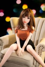 Realistic sex doll with detailed facial features and brown hair, sitting on a beige chair in a cozy indoor setting with bokeh lights in the background. Perfect for adult intimacy and companionship.