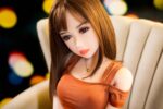 Realistic sex doll female with long brown hair, detailed facial features, wearing an orange top, sitting on a beige chair, perfect for companionship or intimate lifestyle needs.