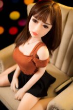 Realistic silicone sex doll with lifelike features, brown hair, and detailed facial expression sitting on a beige sofa. Perfect for companionship and adult entertainment. High-quality craftsmanship.