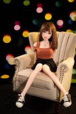 Realistic sex doll sitting on a beige armchair, wearing a brown top and black skirt, with a blurred colorful bokeh background, designed for companionship and adult intimacy.