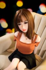 Realistic sex doll with brown hair, designed for adult intimacy, sitting on a beige sofa, with a blurred colorful background, highlighting high-quality craftsmanship and lifelike features for enhanced pleasure.