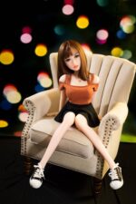 Realistic sex doll female with brown hair, seated on a beige armchair, wearing an orange off-shoulder top, black skirt, and sneakers, with a blurred colorful background.
