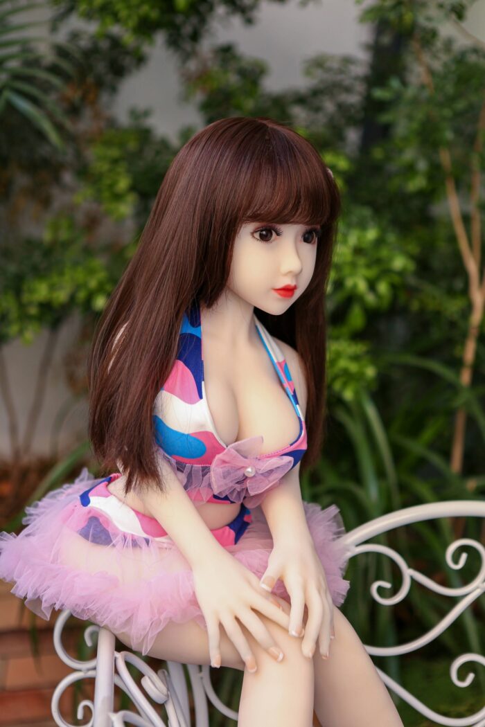 Silicone Sex Doll With Realistic Features, Long Brown Hair, And Expressive Face, Seated Outdoors On A Decorative Metal Bench Amidst Lush Greenery. Perfect For Companionship And Intimate Experiences.