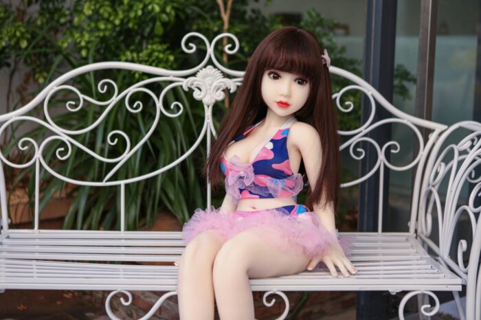 Silicone Sex Doll Sitting On A Stylish White Garden Bench With Lush Greenery In The Background, Featuring Realistic Facial Features, Long Brown Hair, And A Colorful Pink And Blue Outfit.