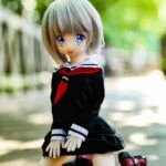 Fluffy anime-style doll with large blue eyes and gray hair wearing a Japanese school uniform sitting outdoors on a wooden deck in sunlight.