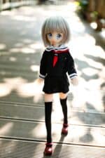 Nukke, jolla on anime-tyyliset kasvonpiirteet, lyhyet hopeanväriset hiukset, yllään musta koulupuku, jossa on valkoinen kaulus ja punainen solmio, kävelee ulkona puisilla lankuilla.