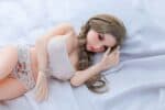 Realistic sex doll lying on a bed with long brown hair, detailed facial features, and wearing cute lingerie, designed for adult pleasure and comfort, perfect for collectors and enthusiasts.