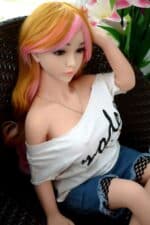Silikon-Sexpuppe mit leuchtend rosa und orangefarbenem Haar, realistischen Gesichtszügen und einem lässigen weißen T-Shirt mit Text. Sie sitzt auf einem Korbstuhl neben rosa Lilien. Perfekt für Intimität unter Erwachsenen und als Sammlerpuppe.
