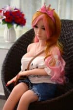 Gigantische Sexpuppe mit rosa und orangefarbenen Haaren, in legerer Kleidung, auf einem Korbstuhl sitzend, realistische Gesichtszüge, lebensgroßes Lächeln, detaillierte Verarbeitung für das Vergnügen Erwachsener, hochwertige Materialien, perfekt für den intimen Gebrauch.