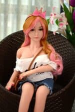 Realistische Sexpuppe mit rosa und orangefarbenem Haar, Freizeitkleidung und rosa Kronen-Stirnband, sitzt in einem Korbstuhl mit Blumenhintergrund. Perfekt für das Vergnügen und die Gesellschaft von Erwachsenen.