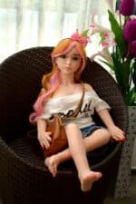 Realistische Sexpuppe mit langen rosa Haaren, Brille und legerer Kleidung, die in einem geflochtenen Stuhl neben Blumen sitzt und für lebensechte Gesellschaft und Vergnügen für Erwachsene konzipiert ist.