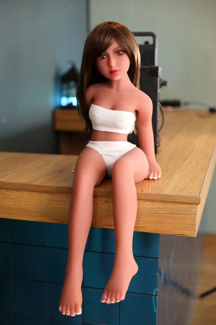 Realistic Sex Doll Sitting On A Wooden Table, Featuring Detailed Features, Long Brown Hair, And A White Crop Top With Matching Bottoms, Designed For Adult Intimacy And Companionship.