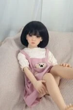 Realistic doll with soft skin, black bob haircut, wearing pink dress with teddy bear pocket, sitting on cozy beige couch for intimate companionship and adult pleasure.