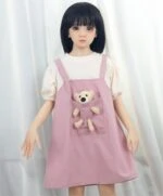 Anime girl doll with black hair in pink dress holding plush teddy bear on a curtain background.