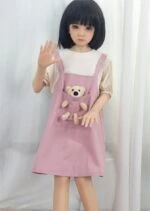 Cute doll girl with black hair in pink dress holding teddy bear, standing near white curtain.