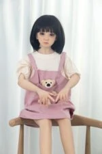 Realistic doll girl with black short hair and big eyes, wearing pink dress with teddy bear pocket, sitting on wooden stool in front of white curtains for exclusive adult comfort and intimacy.