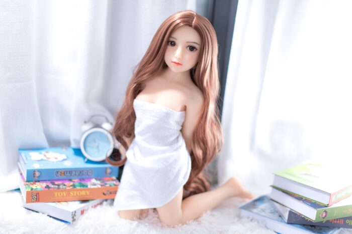 Realistic Love Doll With Long Auburn Hair, Soft Skin, And Detailed Facial Features, Positioned In A Cozy Indoor Setting With Books And A Clock, Emphasizing Lifelike Design And Premium Quality.