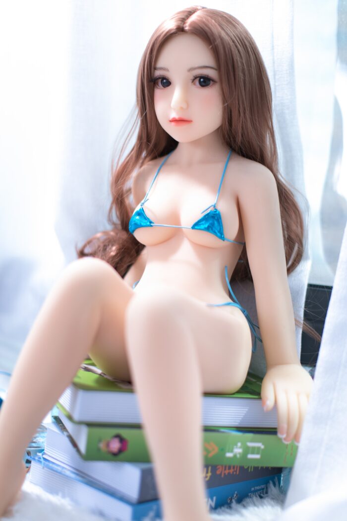 Realistic Sex Doll With Long Brown Hair, Wearing A Small Blue Bikini Top, Posed On Stacked Books Near A Bright Window. Perfect For Experiencing Intimacy And Companionship With High-Quality, Lifelike Features.